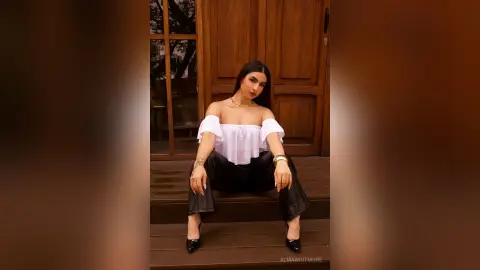AlmaWhitmore sits on wooden steps wearing a white off-the-shoulder top and black pants, looking directly at the camera with long dark hair.