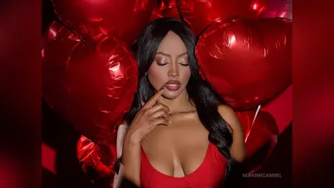 NohomiCambel poses in a red top with her eyes closed and a finger touching her lips, surrounded by red heart-shaped balloons.