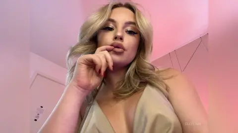 Blonde woman in a gold satin dress posing with a soft, glamorous look against a pink background.