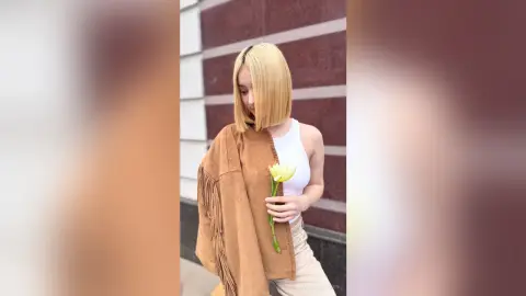 Blonde woman in a white tank top and tan trousers holding a yellow flower against a modern building.