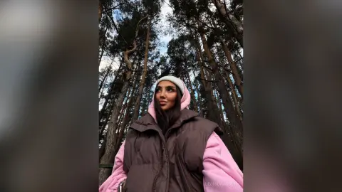 LeonidaAbdon wearing a pink hoodie and a brown puffer vest, looking up while standing in a forest of tall trees.