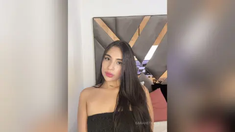 Young woman with long dark hair wearing a black strapless top in a bright modern bedroom.