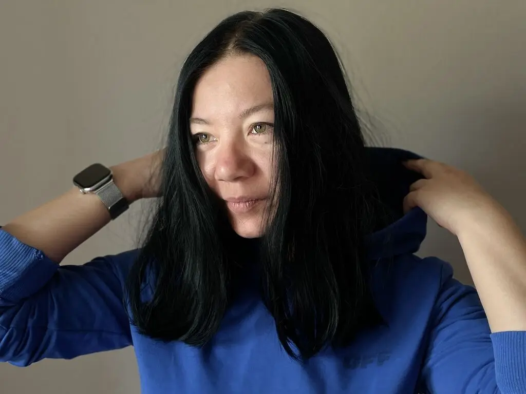 Smiling woman with long black hair wearing a blue hoodie in a minimalist indoor setting.