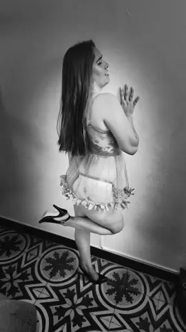 Black and white portrait of a woman in a sheer lace nightgown posing against a wall in a room with patterned tiles.