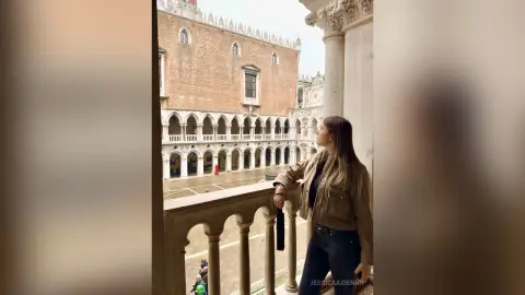 JessicaAidennn — Woman in a tan jacket looking at historic Venetian architecture from a stone balcony on a cloudy day.