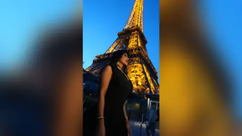 Dolce poses in a black sleeveless dress against the lit Eiffel Tower at night.