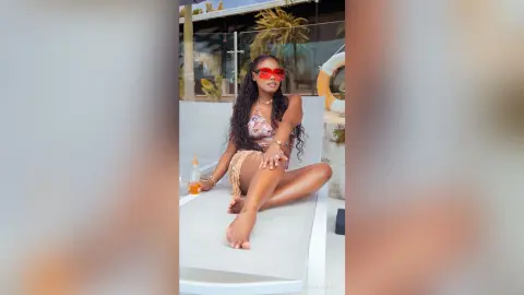 Stylish woman in floral bikini and red sunglasses relaxing on a lounge chair at a sunny tropical resort.