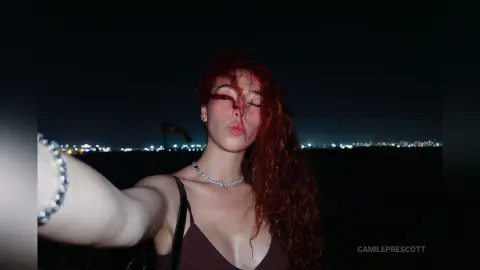 Red-haired woman in a dark top taking a nighttime selfie against a glowing city skyline background.
