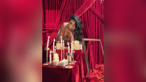 Glamorous woman in gold outfit posing by lit candles against red velvet curtains in a moody, romantic setting.