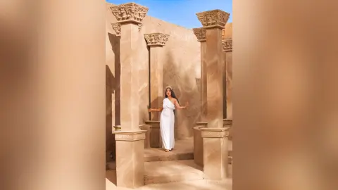 JuliahLewis stands between tall stone pillars wearing a long white dress against a tan wall under a clear blue sky.
