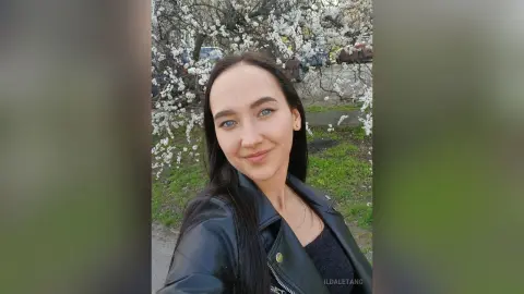 Smiling young woman in a black leather jacket standing before blooming white cherry blossom trees.