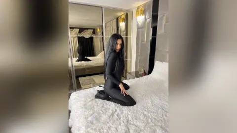 Elegant woman in a black long-sleeve bodysuit sitting on a white faux fur bed in a modern luxury bedroom.