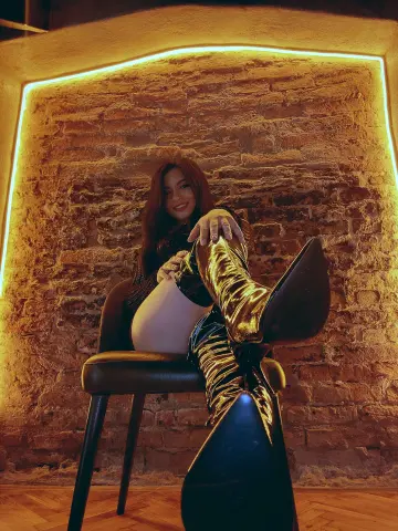 Smiling woman in black outfit and gold metallic boots sitting on a chair against a warm brick wall.
