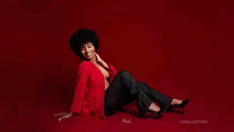 Smiling woman in a red blazer and black trousers sitting against a vibrant red background.