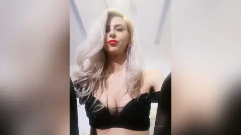 Glamorous blonde woman in a black lace corset top posing with a confident expression in a bright room.