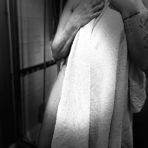 Black and white portrait of a woman wrapped in a white towel in a moody bathroom setting.