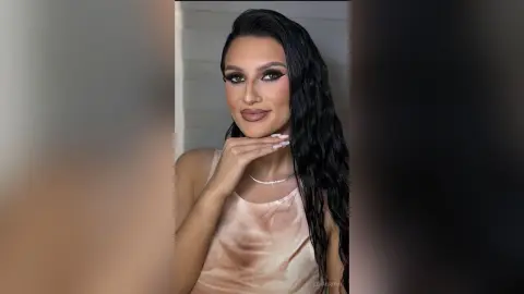 LeyaGrifin poses with long wavy dark hair and heavy eyeliner, wearing a light pink tank top.