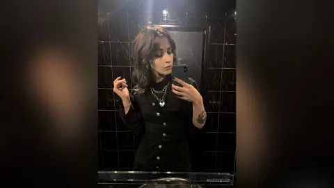 Young woman with wavy hair in a black button-down shirt taking a moody mirror selfie in a dark tiled bathroom.