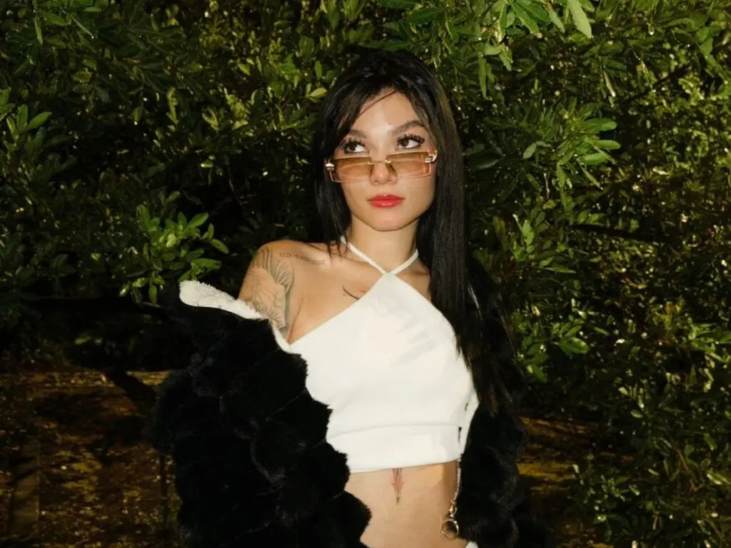 Young woman in a white halter top and black faux fur coat posing against lush green foliage at night.