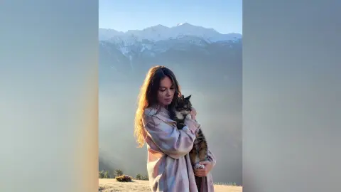 Young woman in a cozy pink plaid robe hugging a tabby cat against a misty mountain landscape at sunrise.