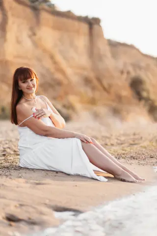 Smiling woman in a white summer dress sitting on a sandy beach near cliffs during golden hour.