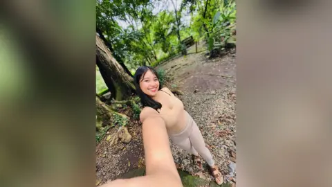 Smiling young woman in a black tank top and beige leggings enjoying a sunny day in a lush green forest.