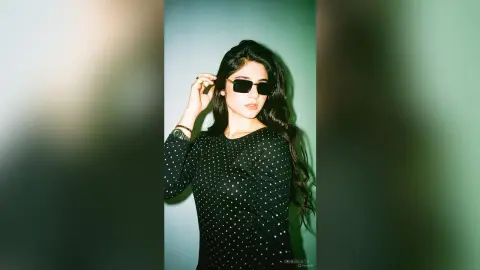 Stylish woman in a black polka dot long-sleeve top and sunglasses posing against a teal studio background.