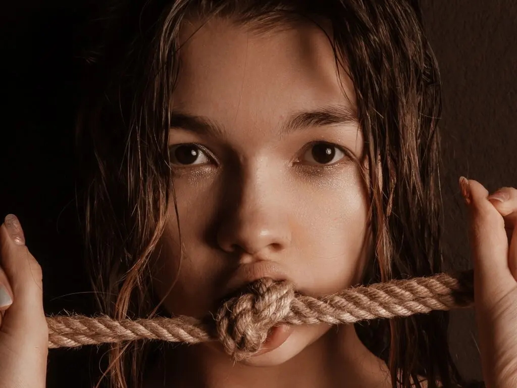 Close-up of a young woman with wet hair holding a thick rope in her mouth in a moody, dark setting.