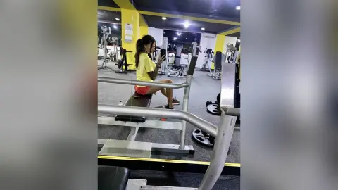 Young woman in yellow gym top sitting on weight machine at fitness center using phone during workout break.