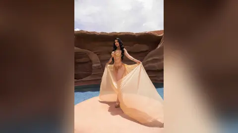 Glamorous woman in a gold bikini and flowing sheer skirt posing against a desert canyon backdrop.