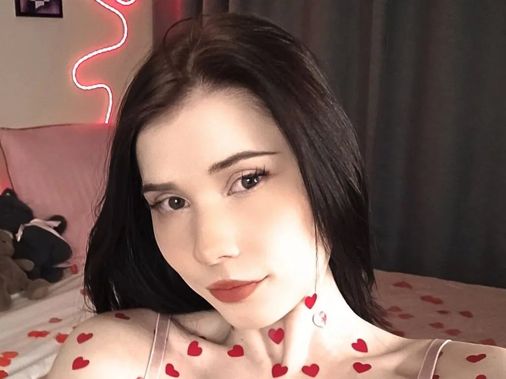 Young woman with dark hair and heart tattoos posing in a cozy room with pink neon lighting.