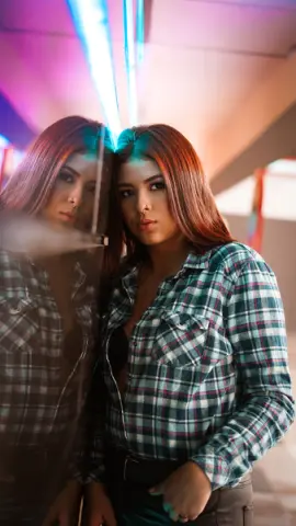 Young woman in a plaid shirt looking into a mirror under moody neon lighting.