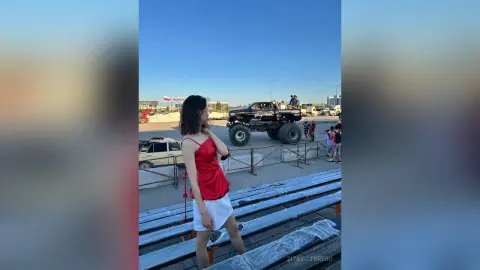 Young woman in a red tank top and white shorts standing outdoors near a large black monster truck.