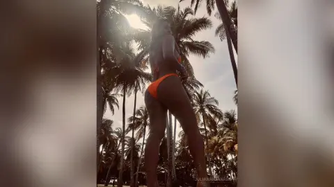 JulianaNavarro poses in an orange bikini, showing her bare back and buttocks while standing outdoors among palm trees.