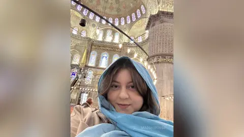 Young woman in a blue headscarf smiling inside the grand, golden Blue Mosque in Istanbul.