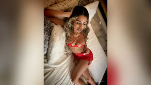 MiaGuerra lies on a white bed wearing a red bra and matching red skirt, running her hands through her long blonde-highlighted hair.