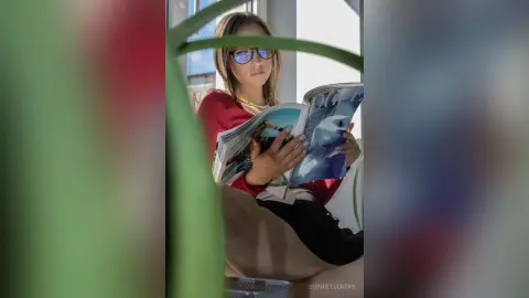 Young woman in red sweater and glasses reading a magazine by a bright window in a peaceful indoor setting.