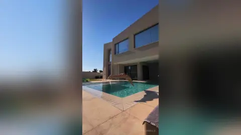 EvelynMills jumps into a clear blue swimming pool next to a modern tan house under a bright blue sky.