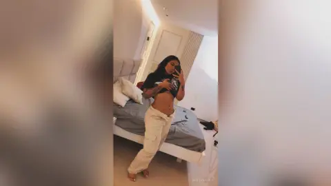Young woman in a black crop top and beige trousers taking a mirror selfie in a bright bedroom.