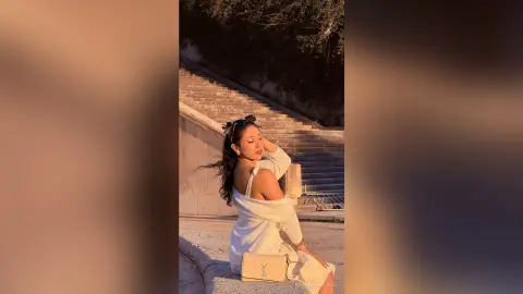 Stylish woman in a white knit sweater sitting on stone stairs during a warm golden hour sunset.