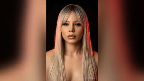 NatashaCastello posing with long blonde hair and heavy makeup against a dark background.
