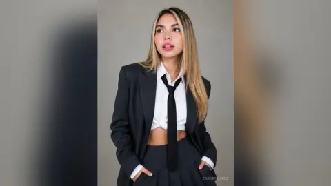 Young woman in a professional black blazer and white shirt with a black tie posing against a gray background.