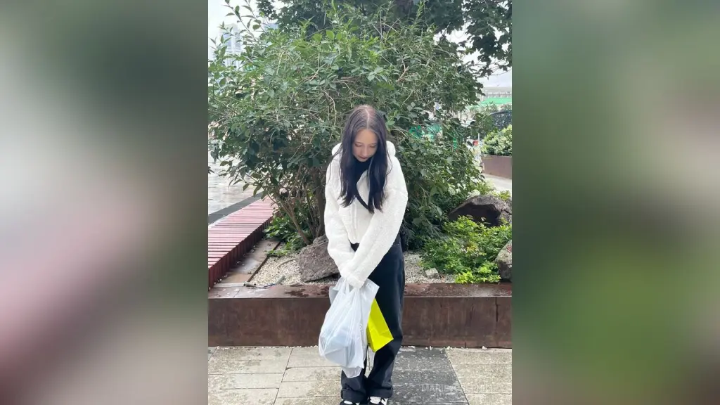 MarieJosePhilip β MarieJosePhilip stands outdoors in front of green bushes, wearing a white long-sleeve top and black pants while holding a white plastic bag.