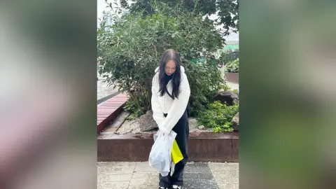 MarieJosePhilip stands outdoors in front of green bushes, wearing a white long-sleeve top and black pants while holding a white plastic bag.