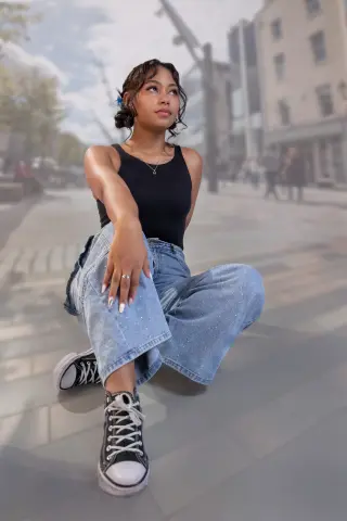Young woman in black tank top and baggy jeans sitting on a city street in casual urban style.