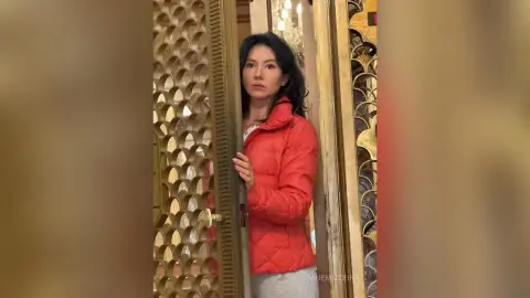 MnemoZeina stands in a doorway wearing a red puffer jacket and light grey pants, looking toward the camera with long dark hair.