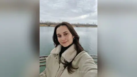 LateshaKaspar smiles while wearing a white fleece-lined jacket, standing outdoors in front of a calm lake and trees.
