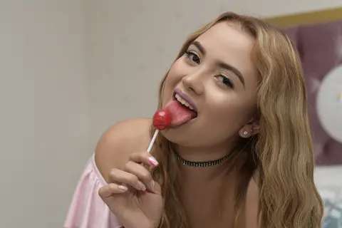 Cheerful young woman with blonde hair licking a red lollipop in a soft, indoor setting.