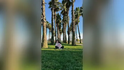 Cheerful woman in a pink sweater sitting on green grass under tall palm trees on a sunny day.