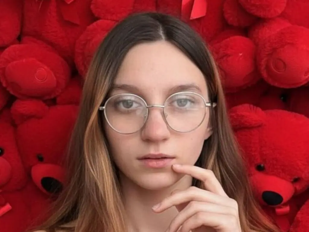 Young woman with glasses posing thoughtfully in front of a wall of red teddy bears.